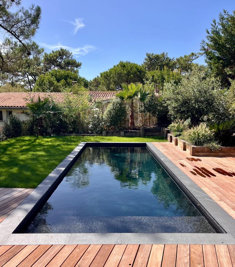 Lien vers la page à propos Piscine rectangulaire aux eaux sombres, entourée d'une terrasse en bois et d'une pelouse verte, avec des arbres en arrière-plan sous un ciel bleu