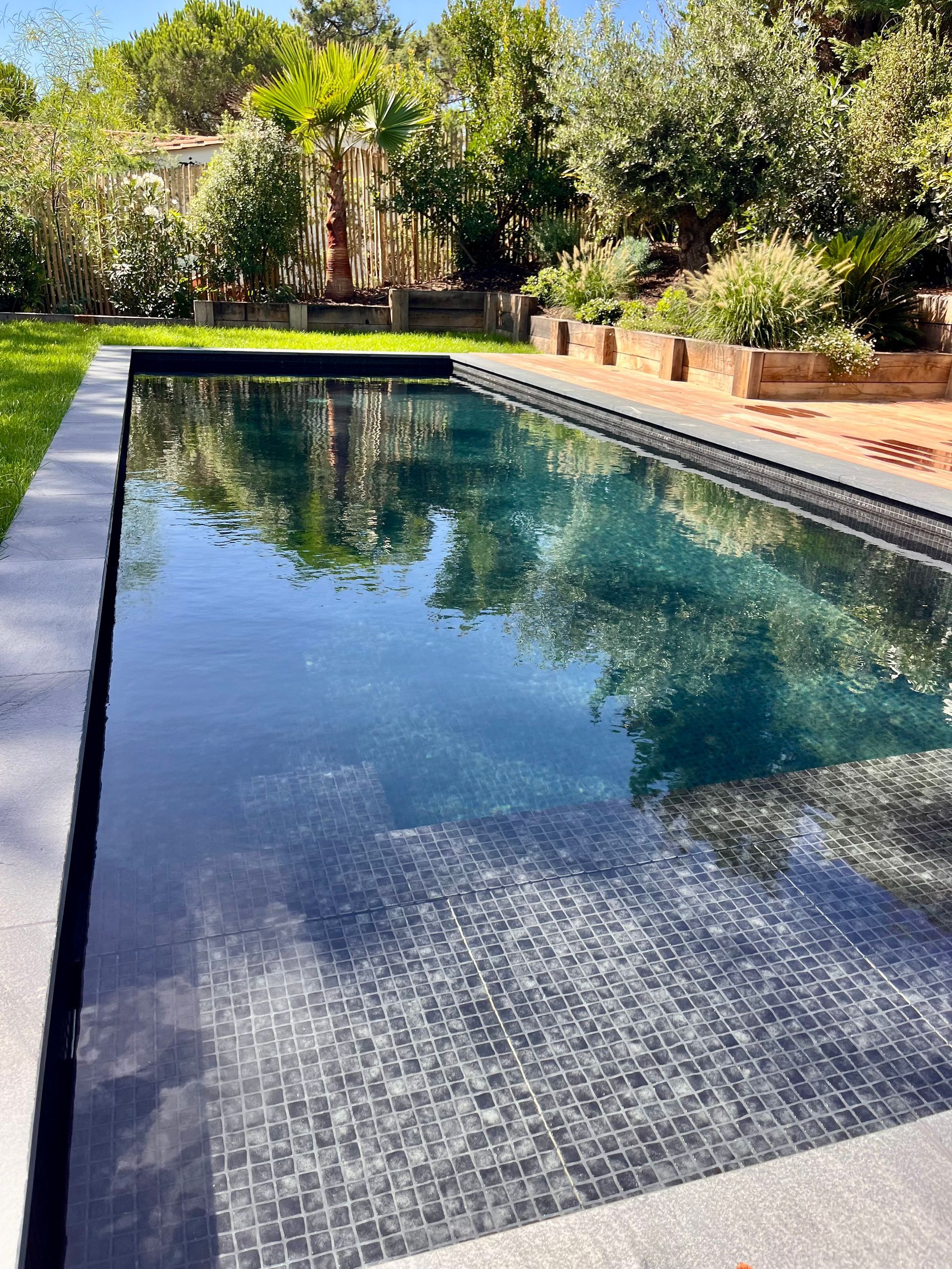 Piscine rectangulaire aux carreaux sombres reflétant le ciel, entourée de pierres et de verdure
