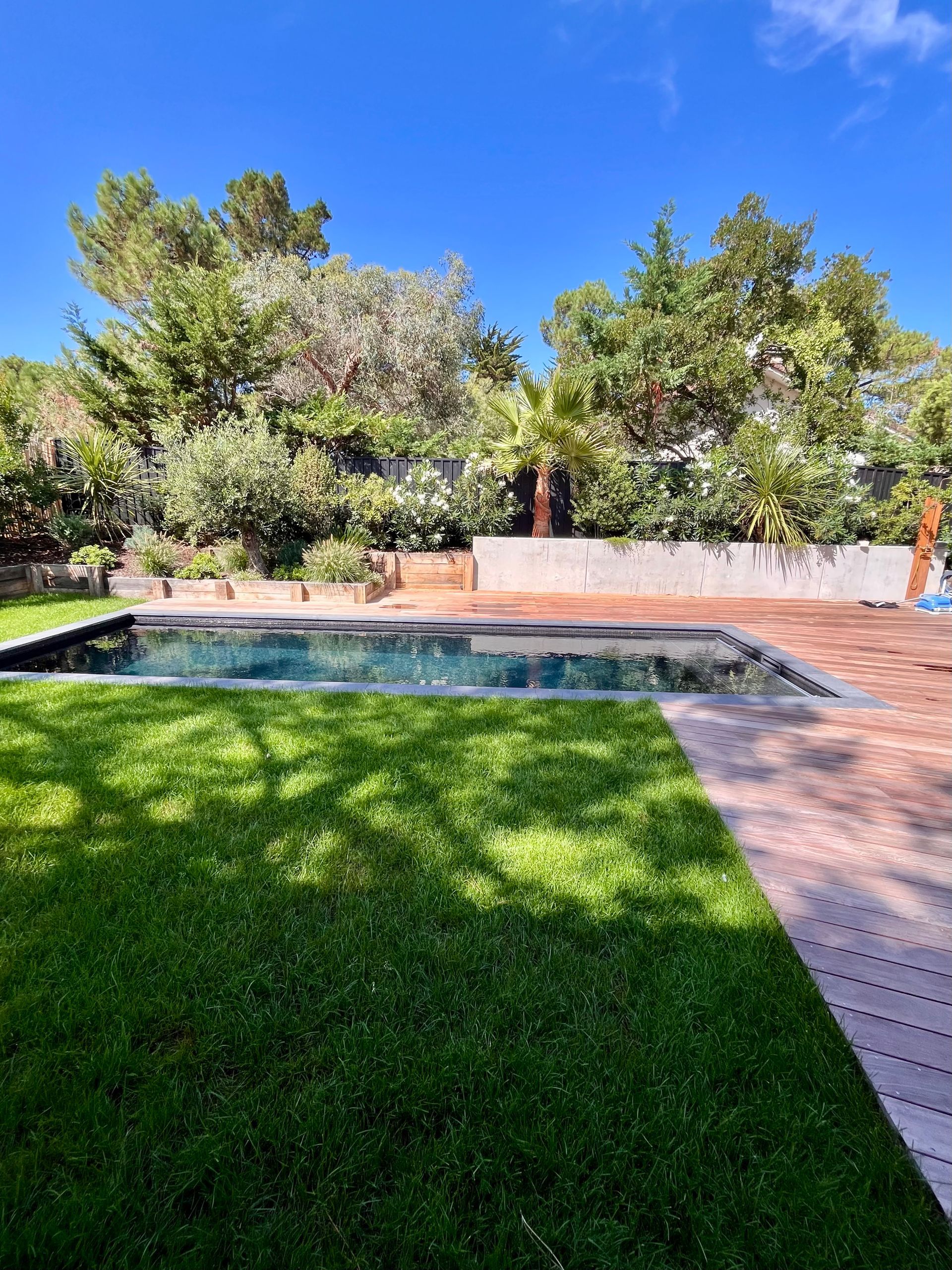 Pelouse verdoyante et piscine avec terrasse en bois sous un ciel bleu, entourée d'arbres