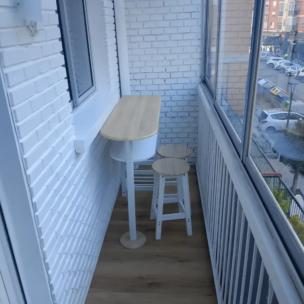 Balcón estrecho con una mesa de bistró blanca y un taburete junto a paredes de ladrillo y ventanas de cristal.