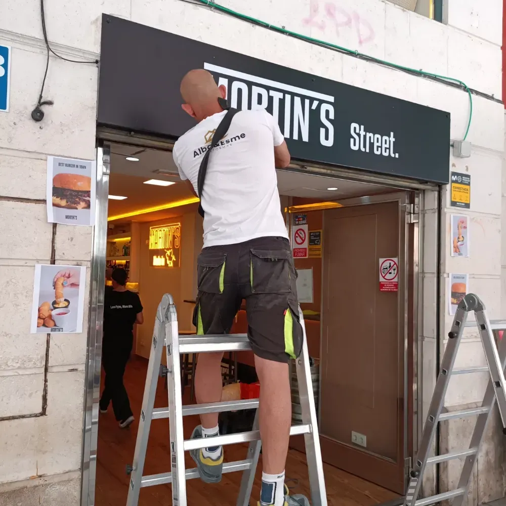 Un trabajador subido a una escalera instala un letrero encima de la entrada de una tienda.