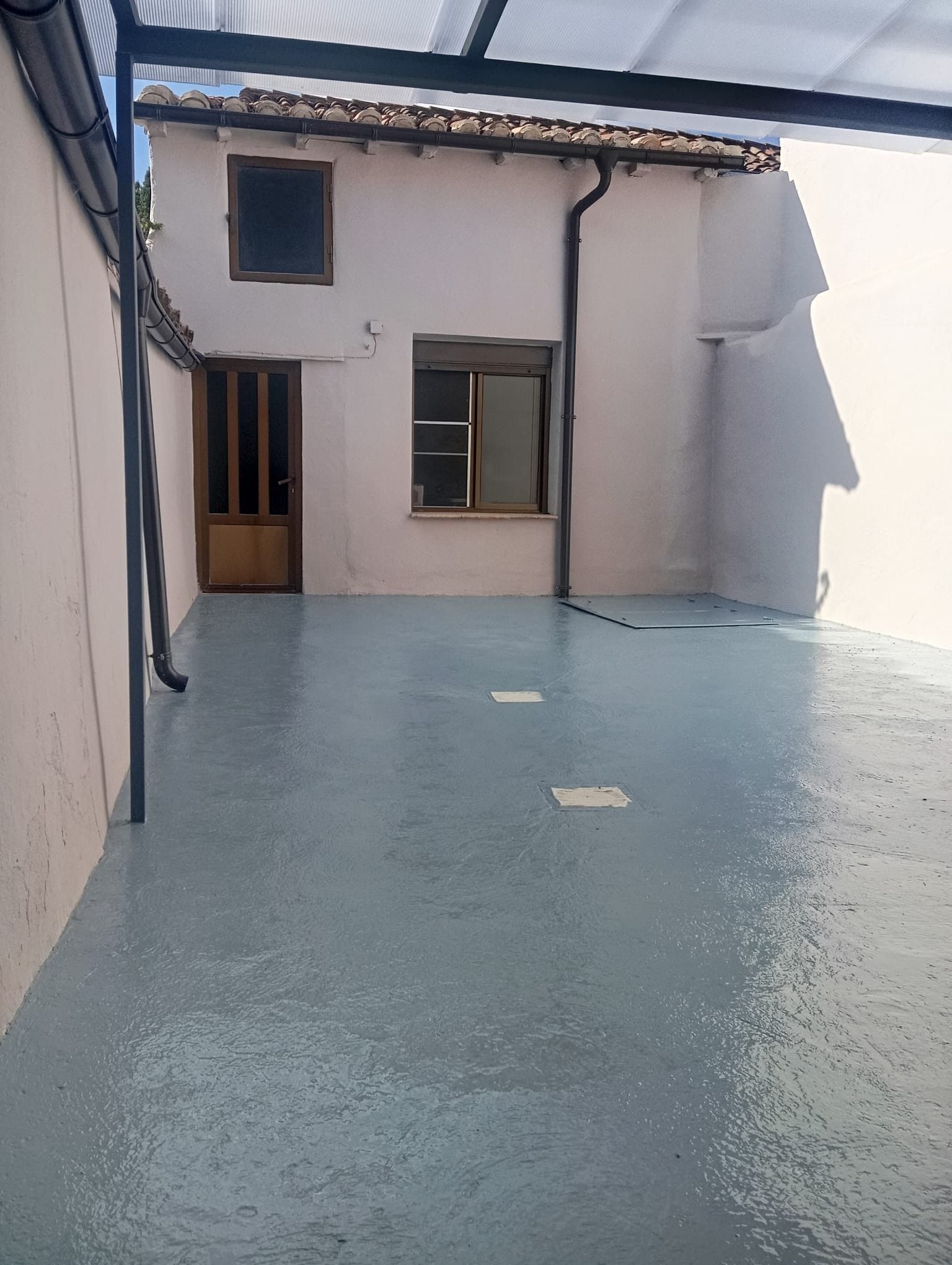 Pequeño patio vacío con suelo azul, paredes blancas y una puerta y una ventana al fondo.