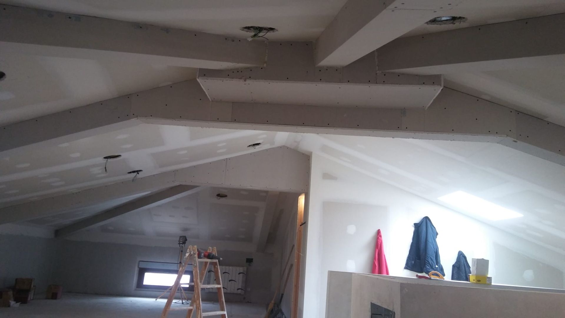 Habitación abuhardillada en obras, con vigas a la vista, una escalera y ropa colgada junto a una ventana luminosa