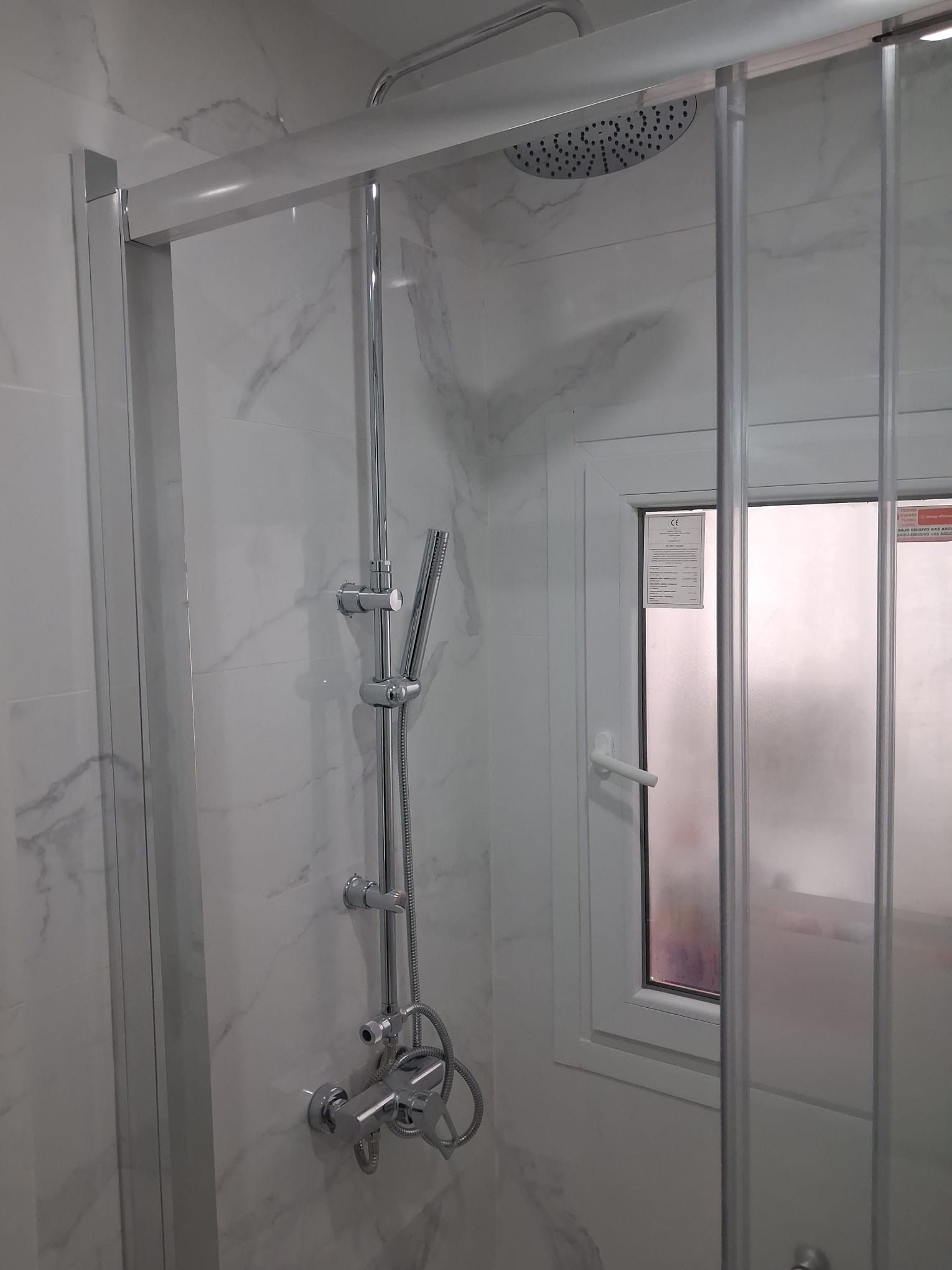Ducha moderna con puertas de cristal, grifería cromada, paredes con efecto mármol y una ventana pequeña