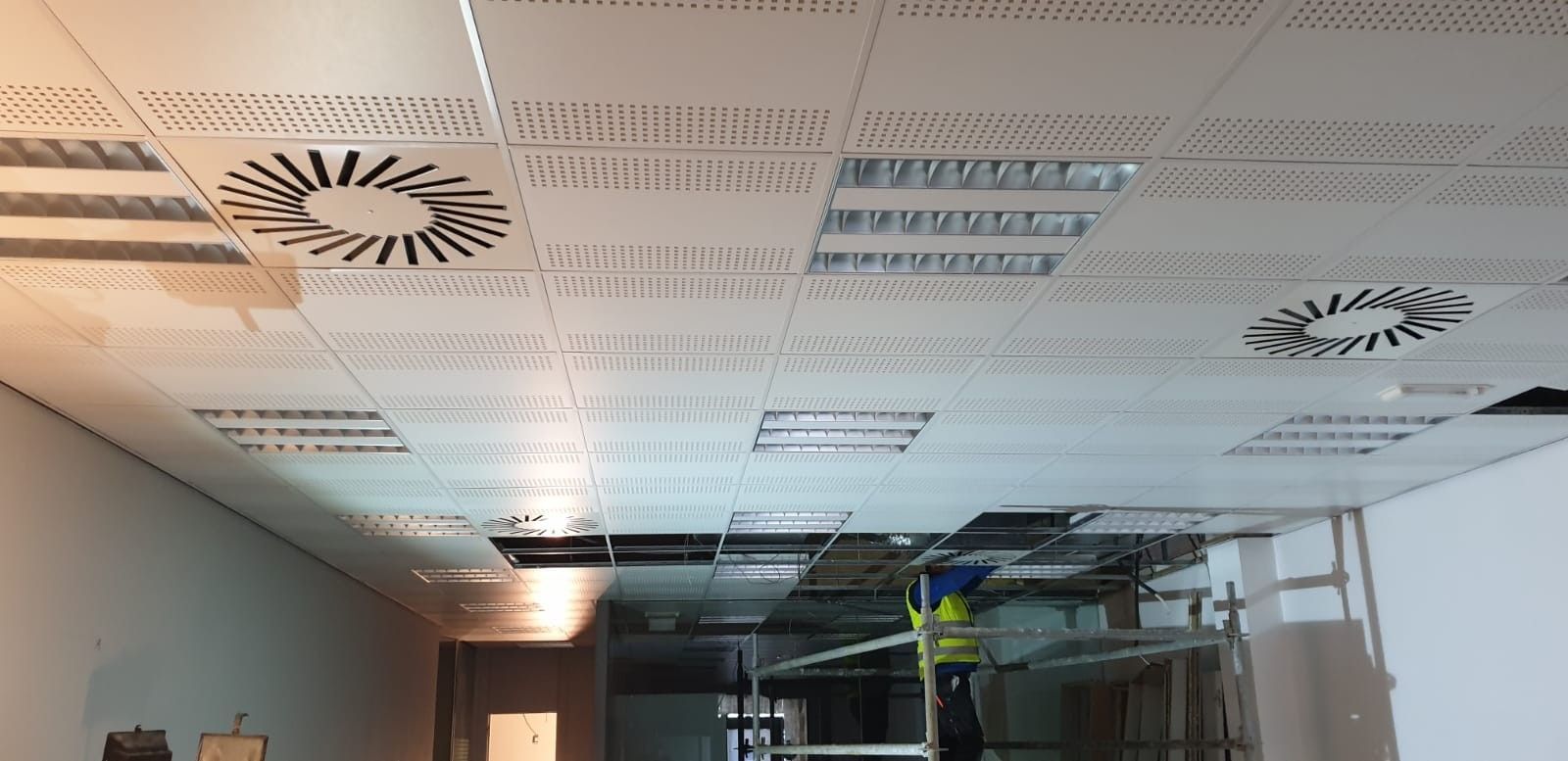 Techo de oficina con placas acústicas, rejillas de ventilación y difusores circulares de aire sobre salas con paredes de vidrio