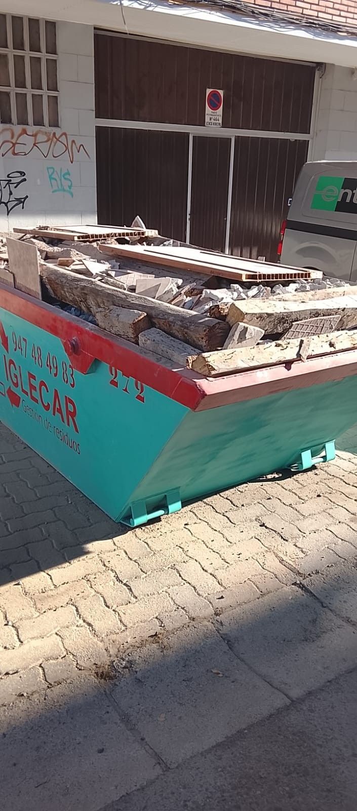 Contenedor verde azulado lleno de escombros de madera sobre una acera de ladrillo frente a un edificio
