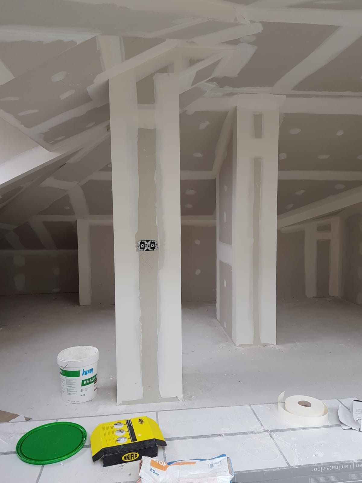 Habitación abuhardillada sin terminar, con paredes de pladur, vigas a la vista y materiales de pintura en el suelo.