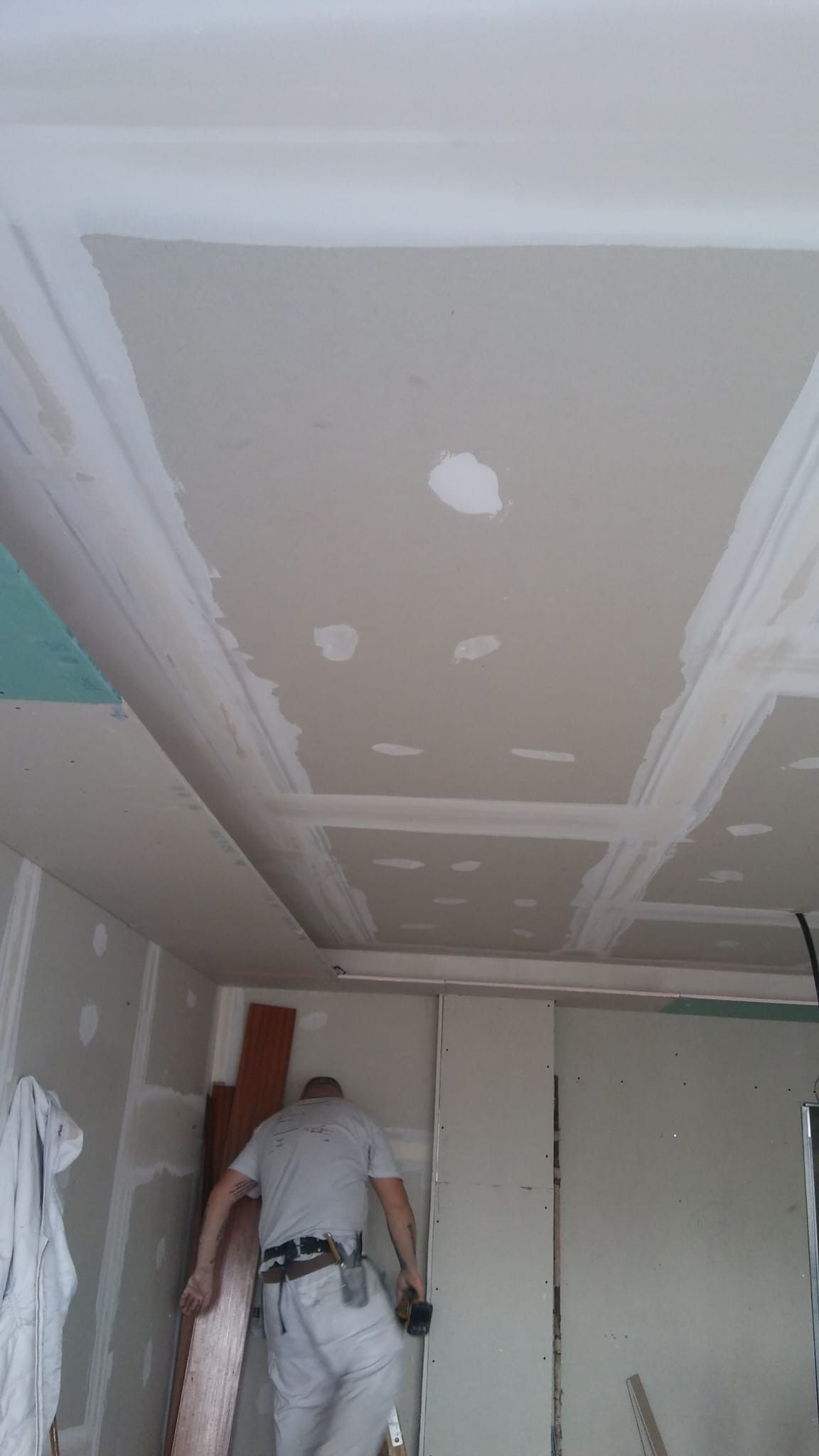 Persona trabajando en el techo de una habitación con parches de paneles de yeso y una escalera, en un entorno de renovación