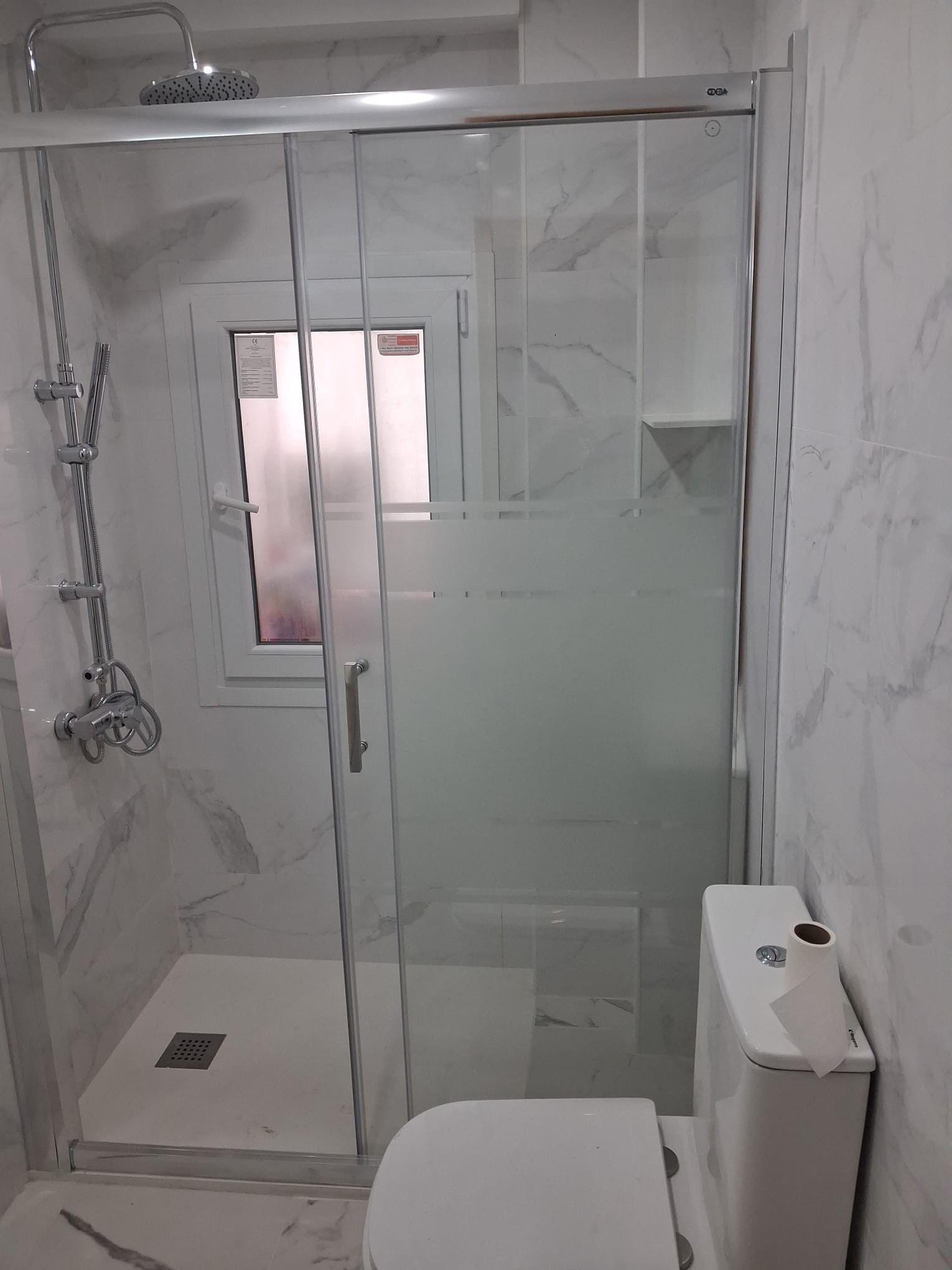 Baño pequeño alicatado con ducha de cristal, puerta corredera esmerilada e inodoro.