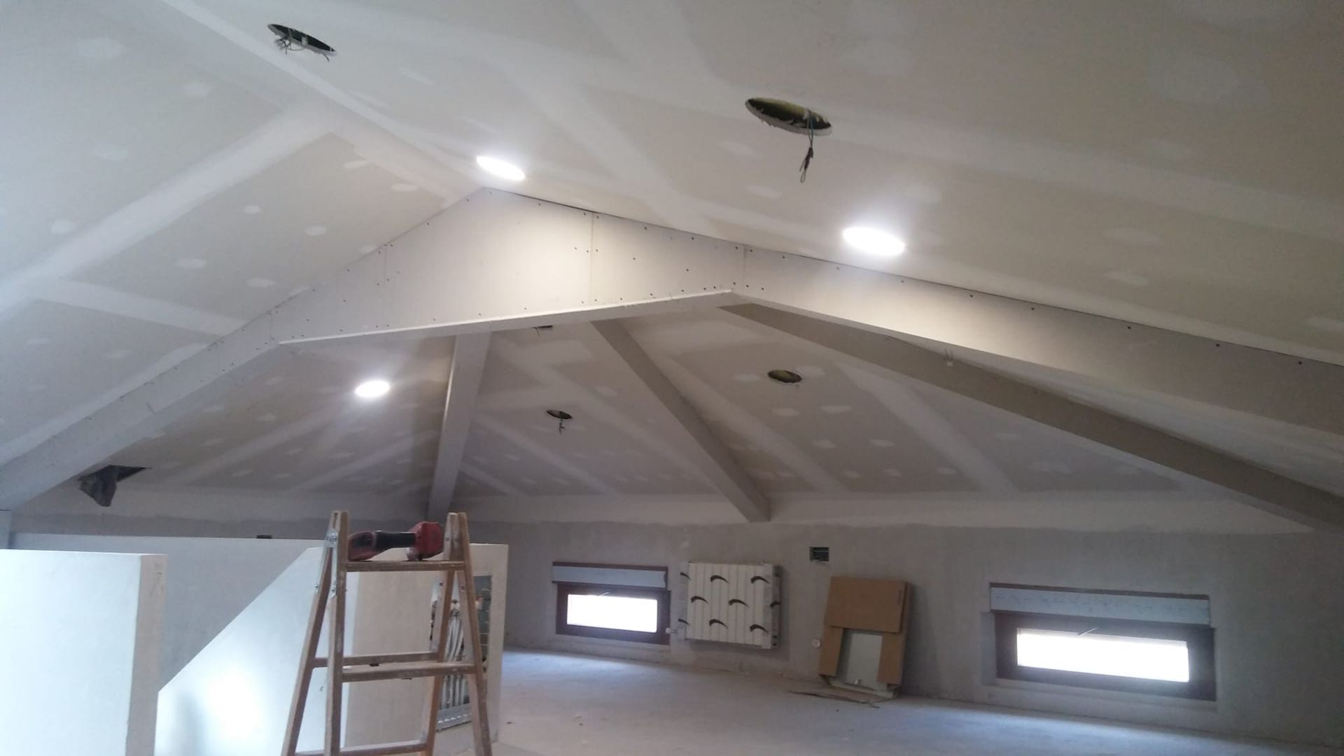 Ático sin terminar, con techo inclinado blanco, luces empotradas, ventanas y una escalera en construcción.