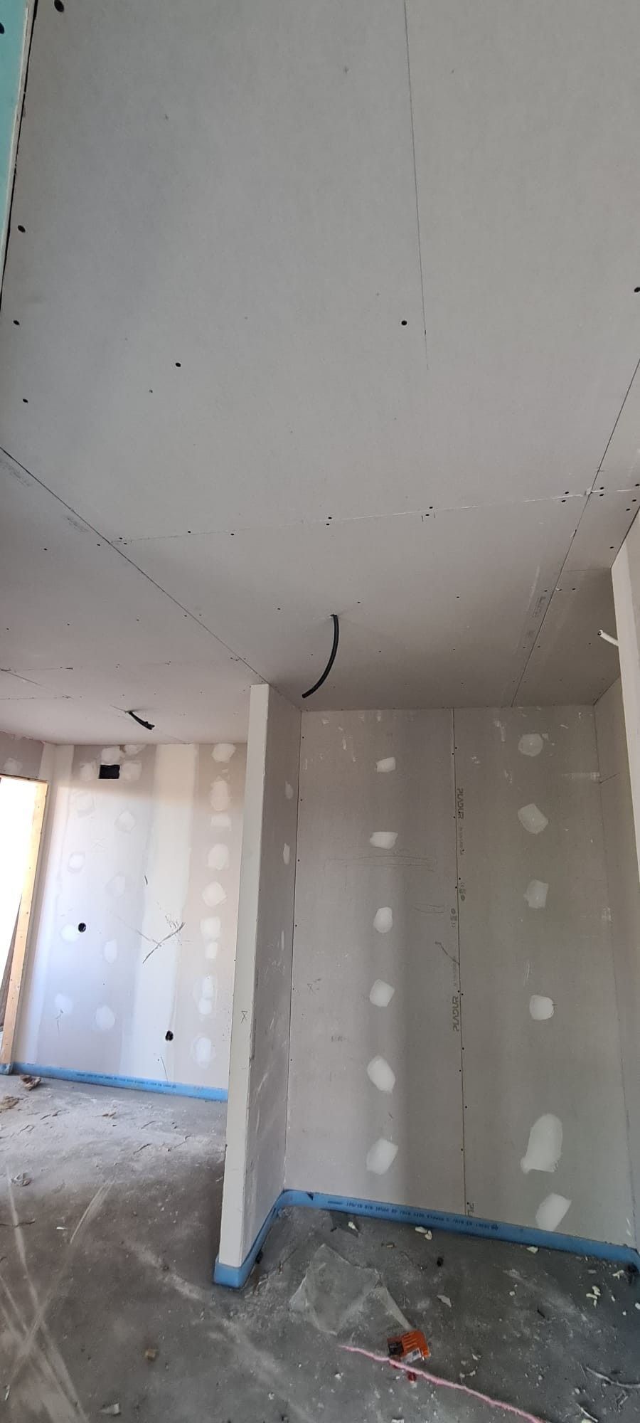 Pasillo interior sin terminar, con paneles de yeso blancos, el techo al descubierto y los bordes del suelo cubiertos con cinta, en obras.