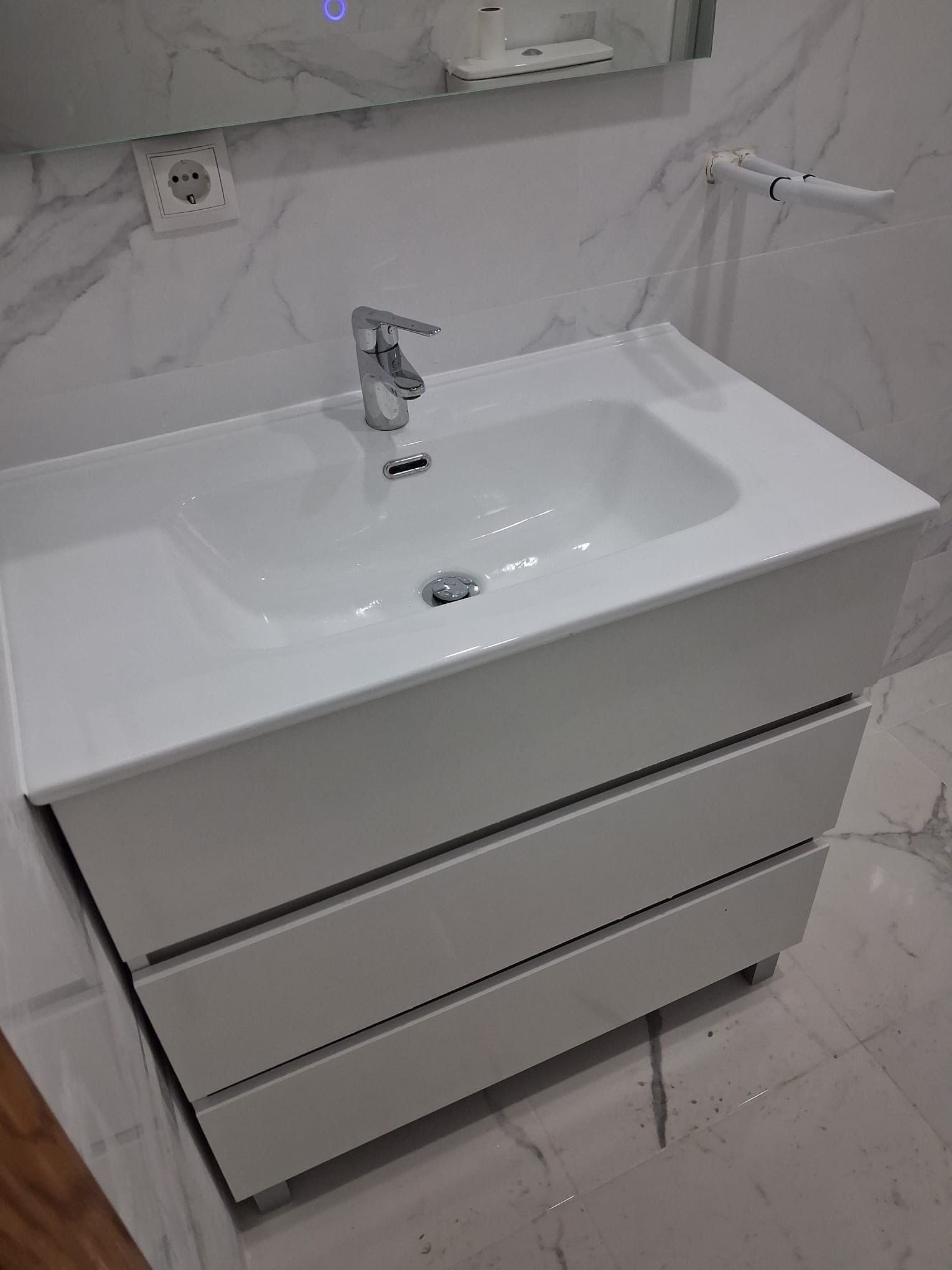 Mueble de baño moderno blanco con lavabo integrado y grifo cromado, bajo una pared con acabado efecto mármol.