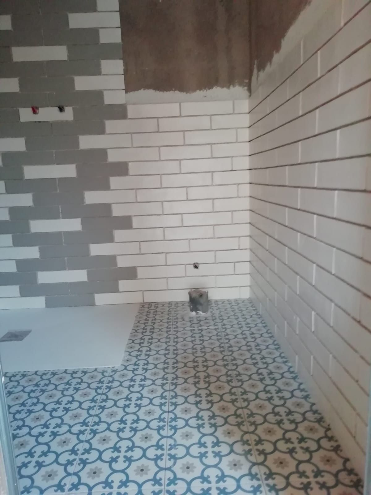 Baño pequeño alicatado en obras, con paredes de azulejos blancos tipo metro y suelo de baldosas estampadas en azul y blanco.