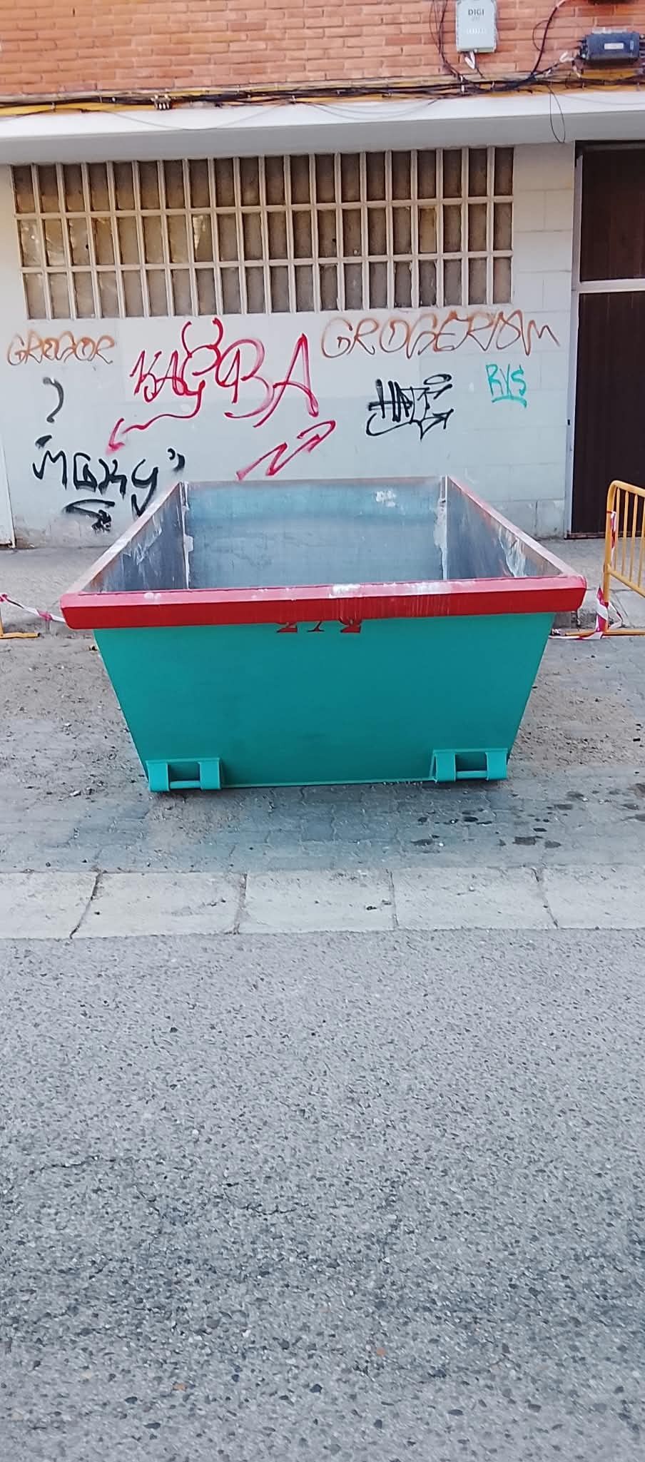 Contenedor grande de color verde azulado con tapa roja en una calle, delante de una pared cubierta de grafitis