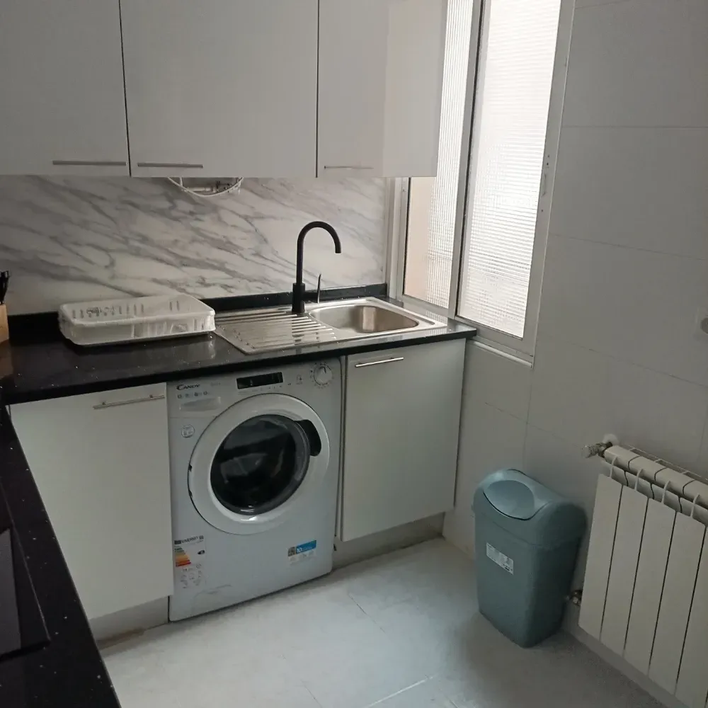 Cocina compacta con encimera negra, fregadero, lavadora, armarios blancos y una ventana junto a un radiador y un cubo de basura.