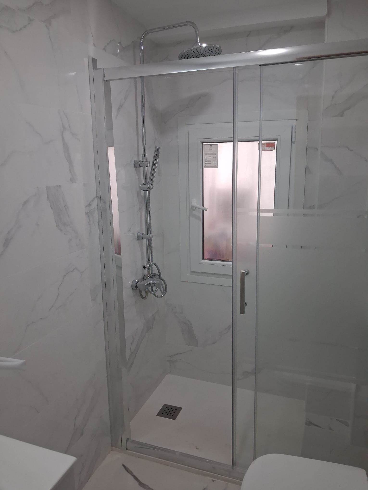 Mampara de ducha de cristal en un baño alicatado, con ducha de mano y desagüe en el suelo.