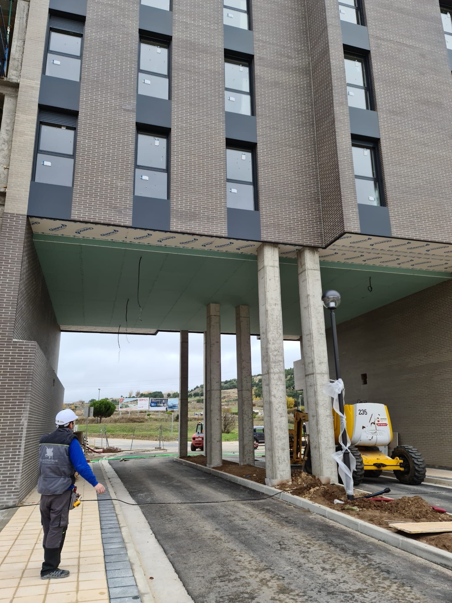 Trabajadores de la construcción junto a la entrada elevada de un edificio, con un camino de acceso pavimentado y columnas de soporte de hormigón