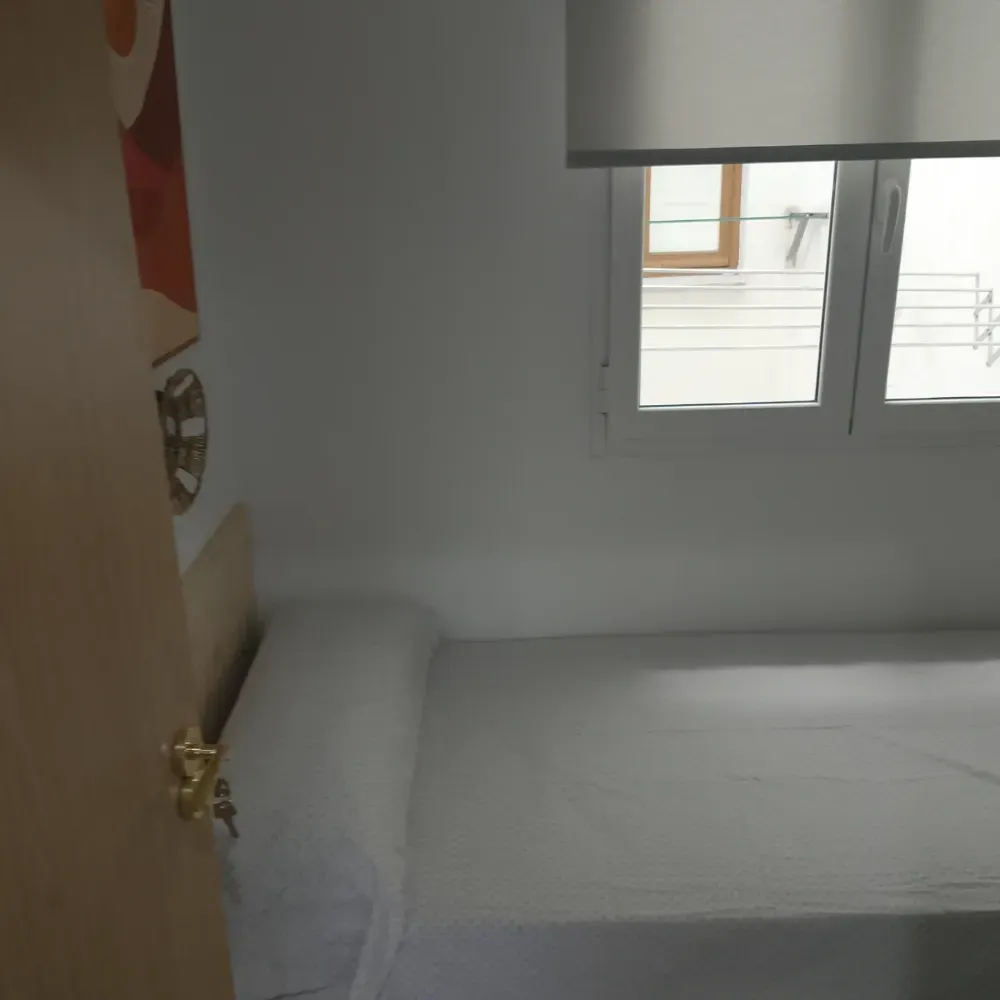 Habitación pequeña con una cama junto a la ventana y una puerta de madera a la izquierda.