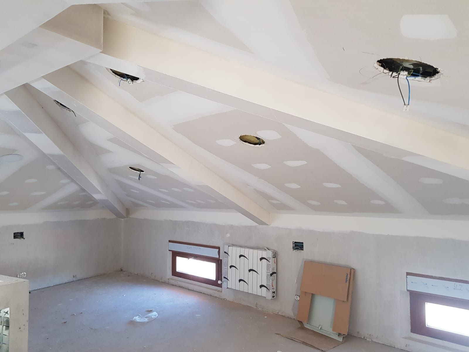 Habitación abuhardillada sin terminar, con techo blanco inclinado, luces a la vista, ventanas y parches de pladur en el suelo.