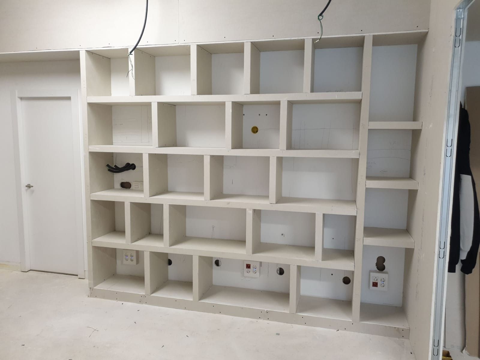 Pared blanca de estanterías empotradas en una habitación en construcción, con varios huecos vacíos y el suelo sin terminar.