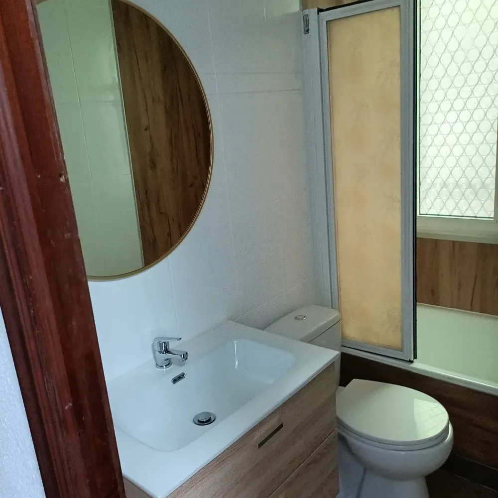 Baño pequeño con lavabo, inodoro, espejo redondo y ventana esmerilada junto a una cabina de ducha.