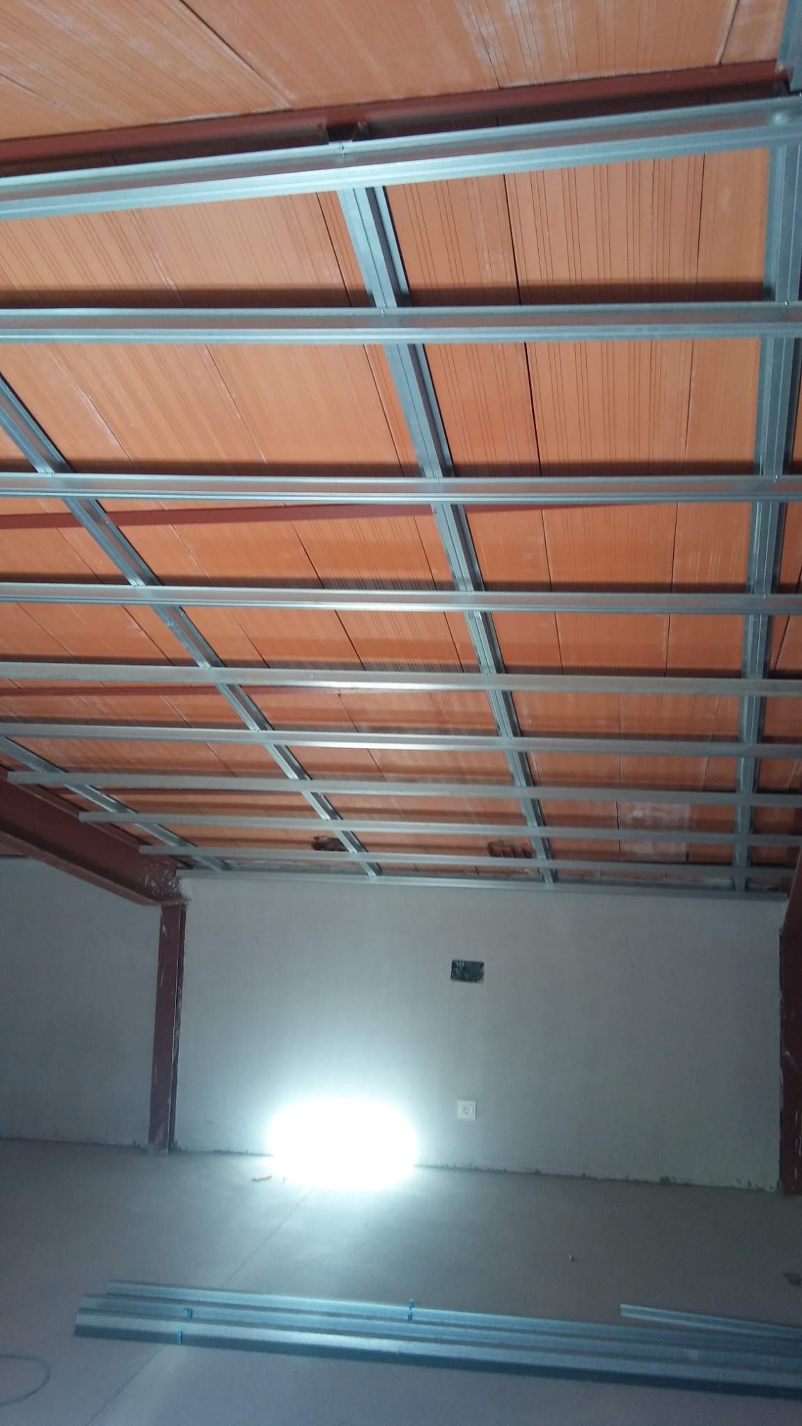 Interior de un garaje vacío con vigas del techo a la vista, una pared blanca y una luz intensa en el suelo, cerca del fondo.