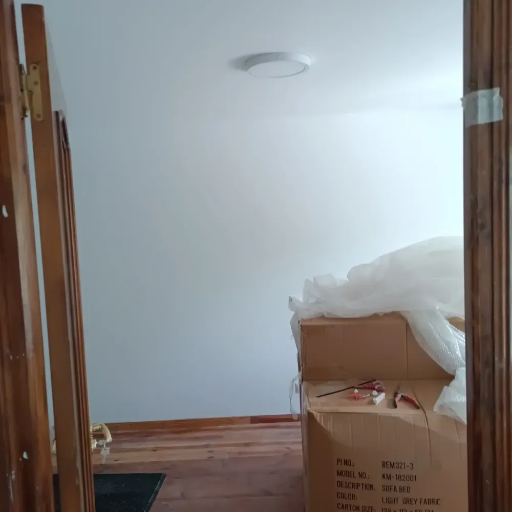 Habitación parcialmente amueblada con paredes blancas, suelo de madera y una caja de cartón cubierta de plástico cerca de la puerta.