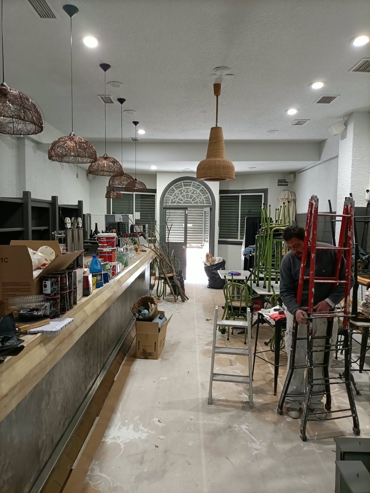 Interior de una tienda o taller desordenado, con lámparas colgantes, mostradores, taburetes y plantas.