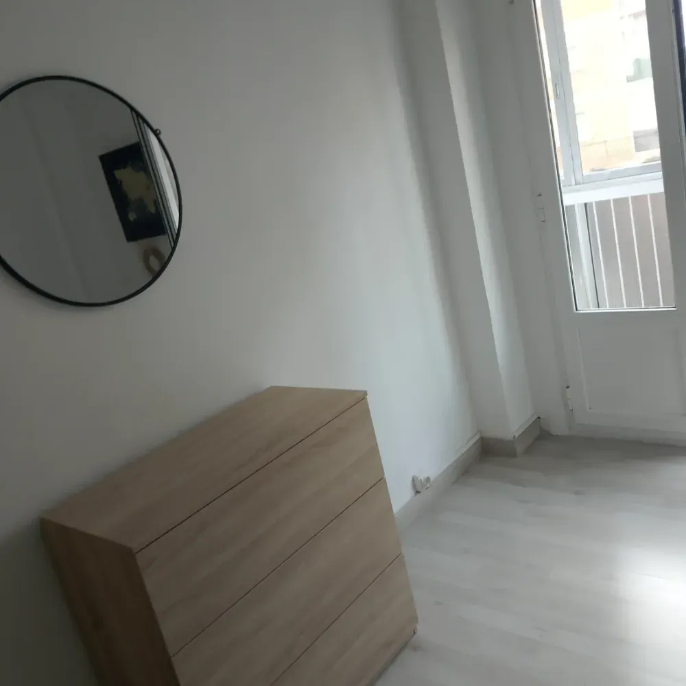 Habitación luminosa y vacía con una cómoda de madera, un espejo redondo en la pared y una puerta que da al balcón.