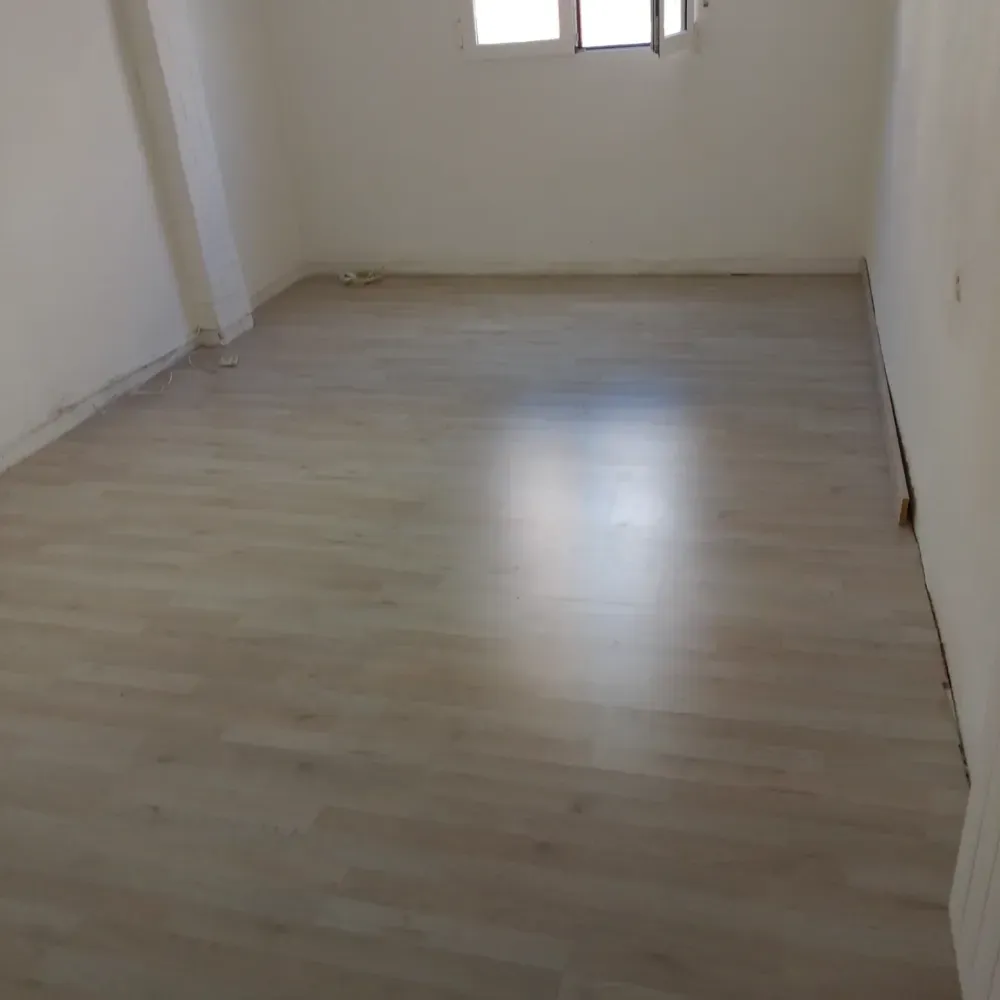 Habitación vacía y luminosa con suelo de madera clara y una pequeña ventana en la pared del fondo.