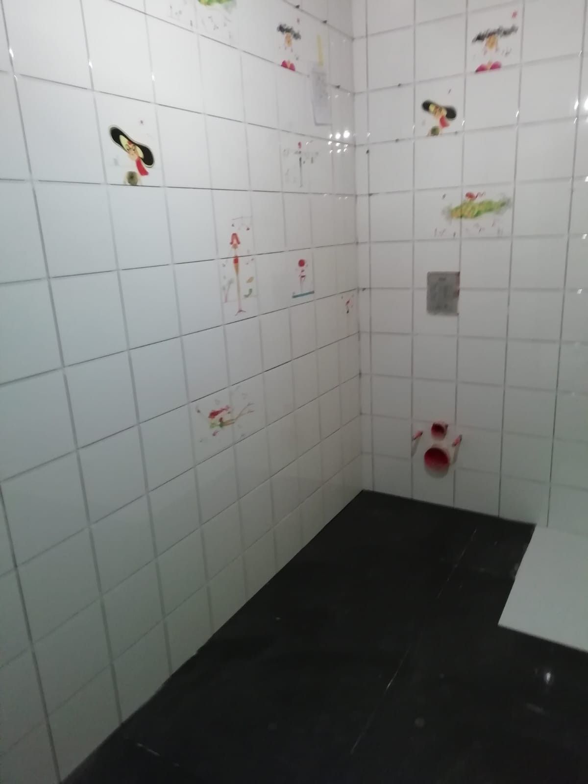 Pequeña habitación con azulejos blancos, suelo negro, paredes marcadas y un desagüe en la esquina.