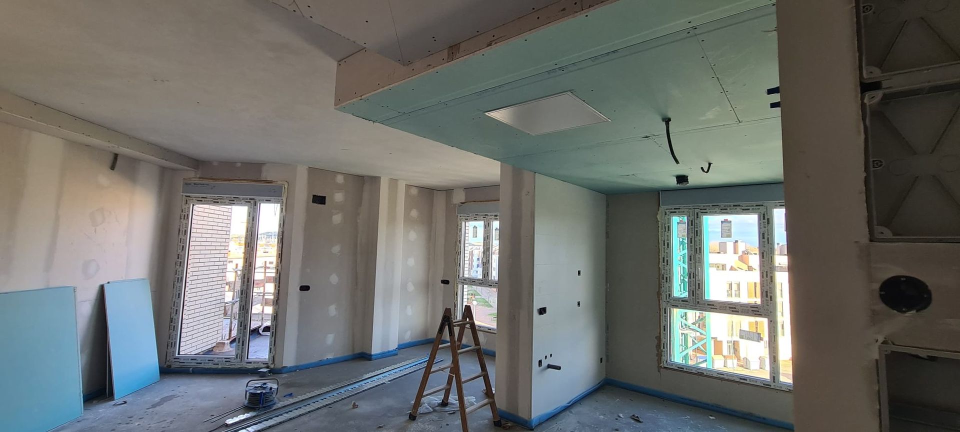 Renovación en curso en una habitación con paneles de yeso a la vista, herramientas y grandes ventanas por las que entra la luz del día