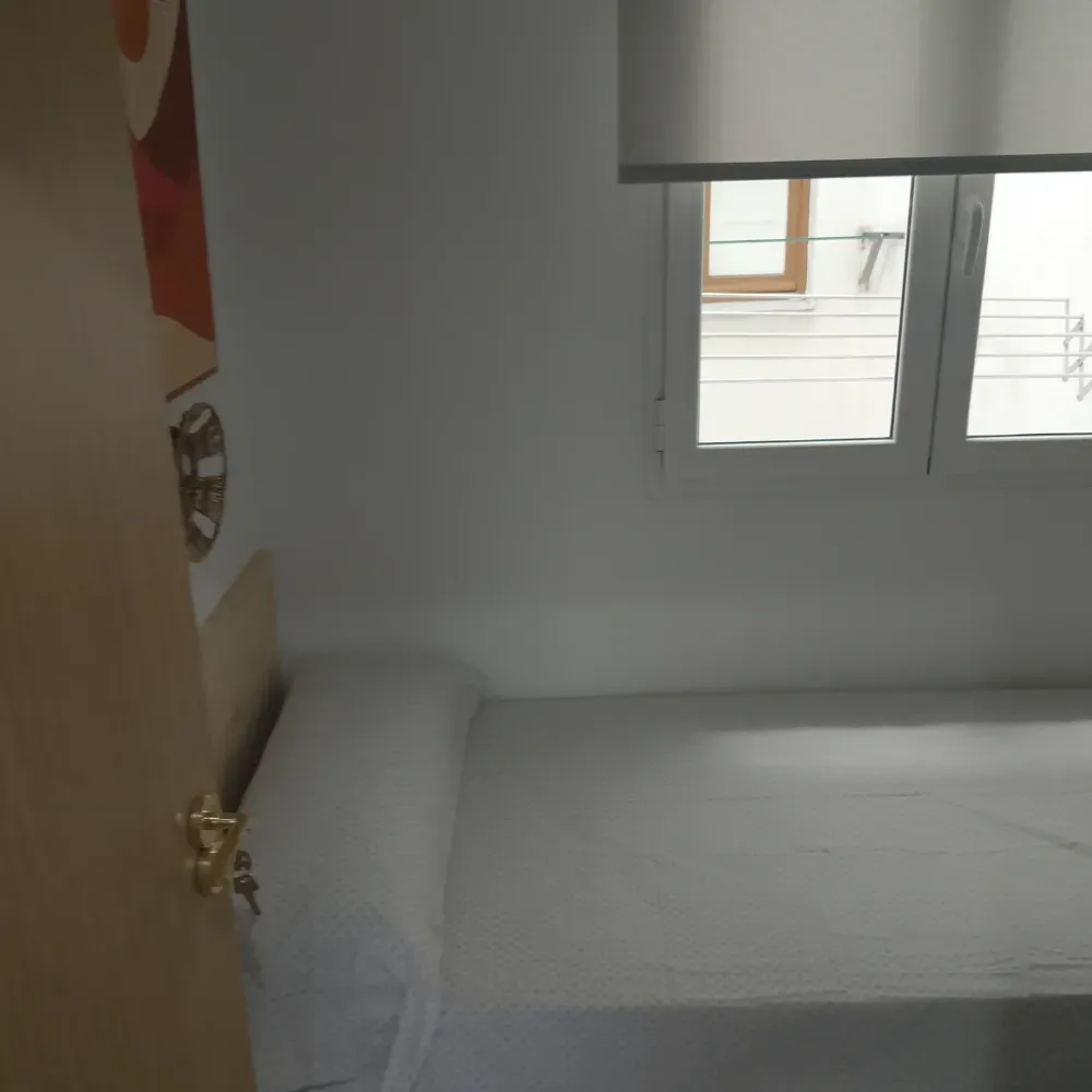 Habitación pequeña con una cama y una almohada junto a una ventana con la persiana parcialmente bajada.