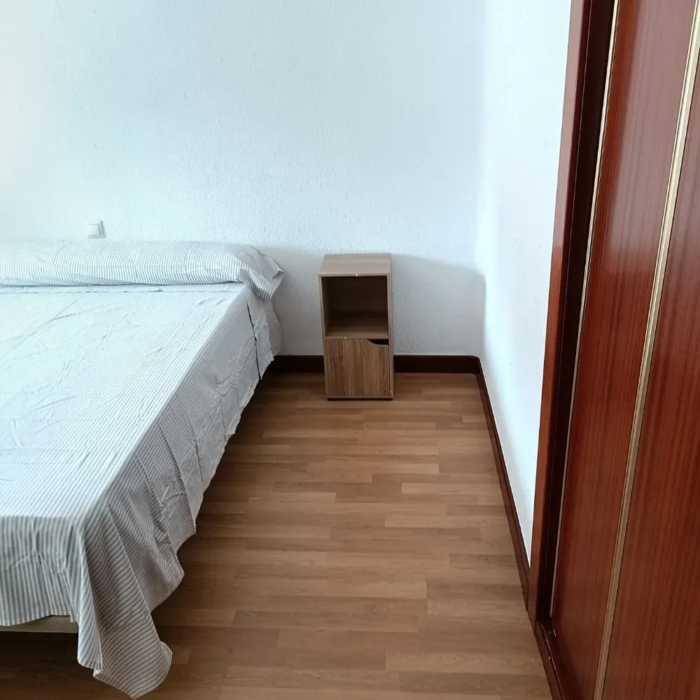 Habitación pequeña con cama, mesita de noche de madera, paredes blancas y suelo de madera.
