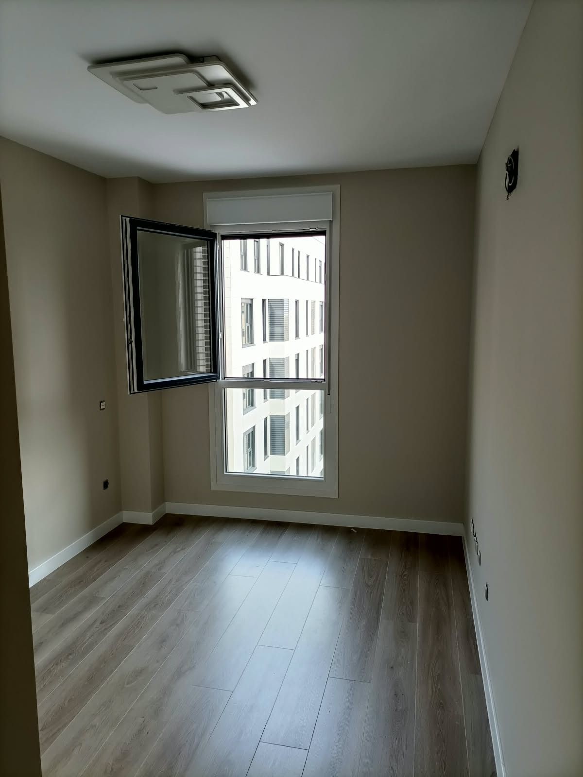 Habitación vacía sin amueblar, con suelo de madera, una ventana abierta y una lámpara de techo en un apartamento.