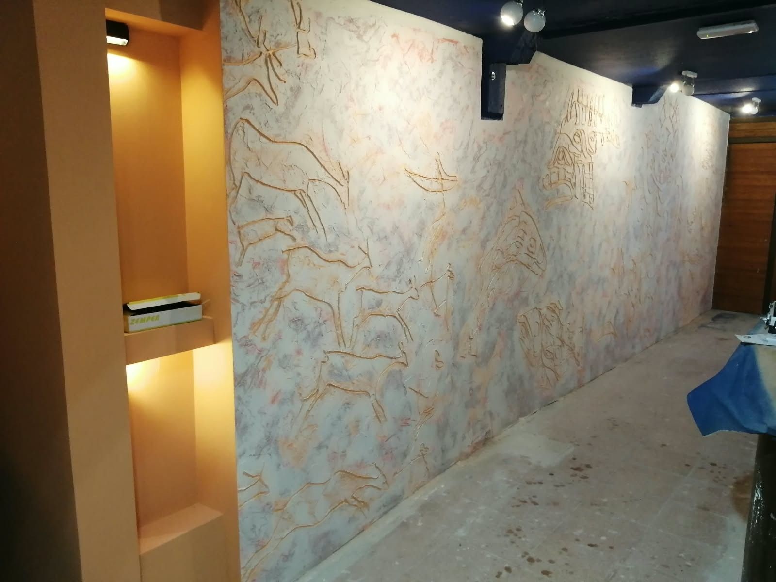 Pasillo vacío con una pared texturizada, iluminación cálida en un nicho de pared y un objeto cubierto de azul a la derecha