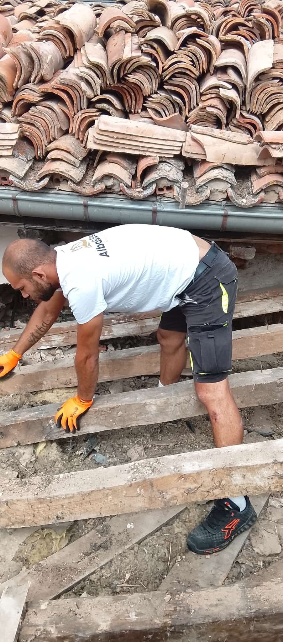 Trabajador con camisa blanca y pantalones cortos negros levantando tejas en un tejado.