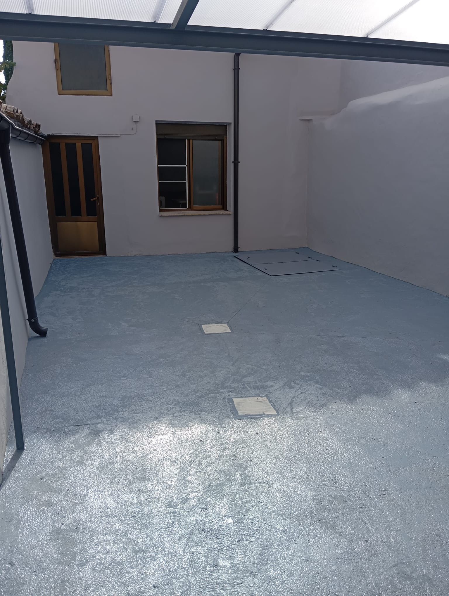 Pequeño patio de cemento vacío, con paredes blancas, dos ventanas y una puerta de madera.