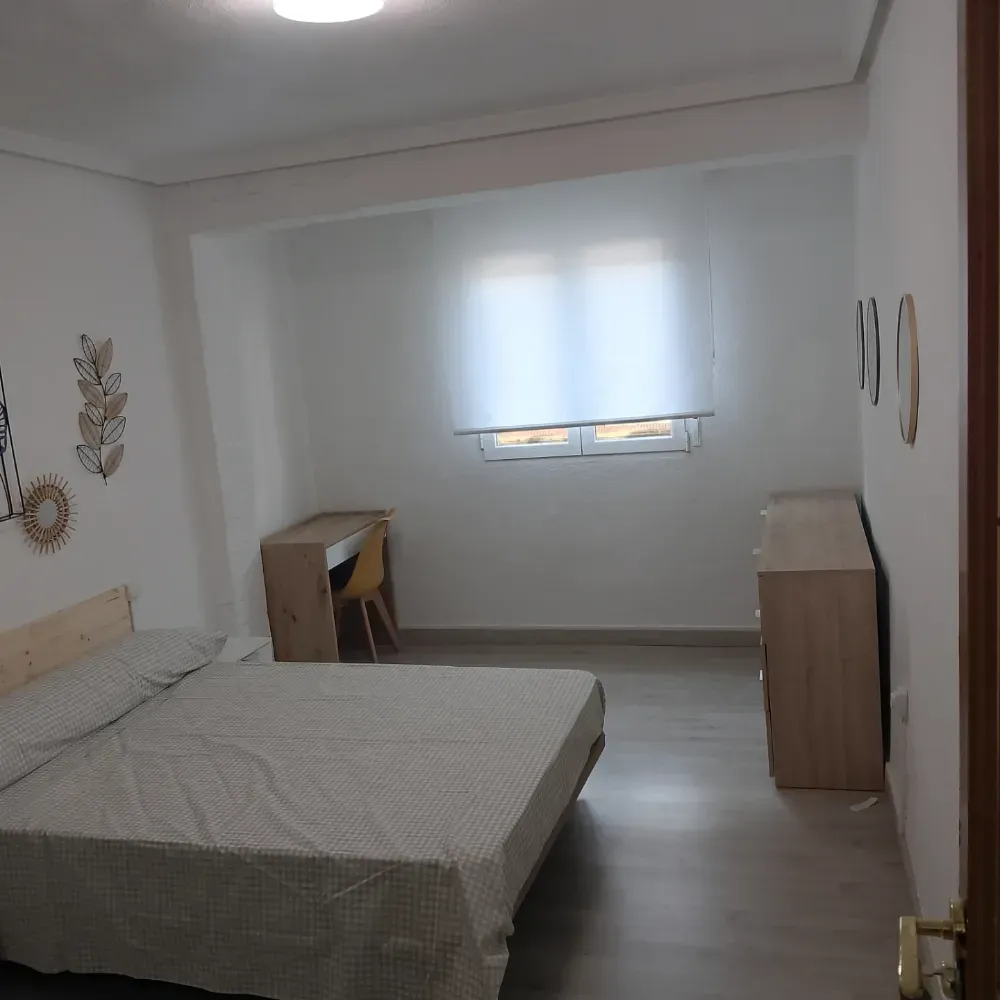 Dormitorio minimalista con cama, escritorio, cómoda y ventana con persiana blanca.