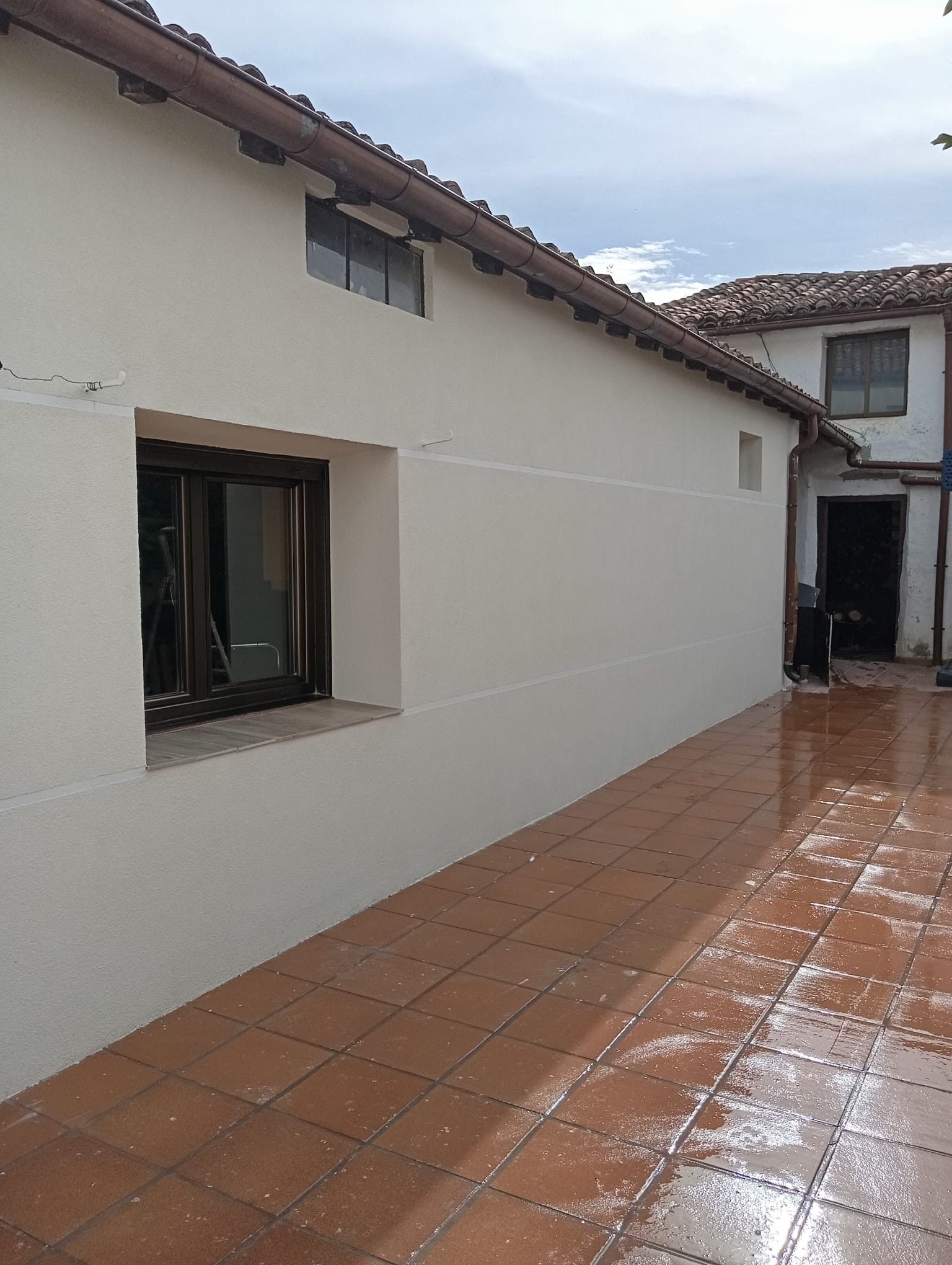 Patio alicatado mojado junto a una casa blanca con una ventana y tejado de tejas, bajo un cielo nublado