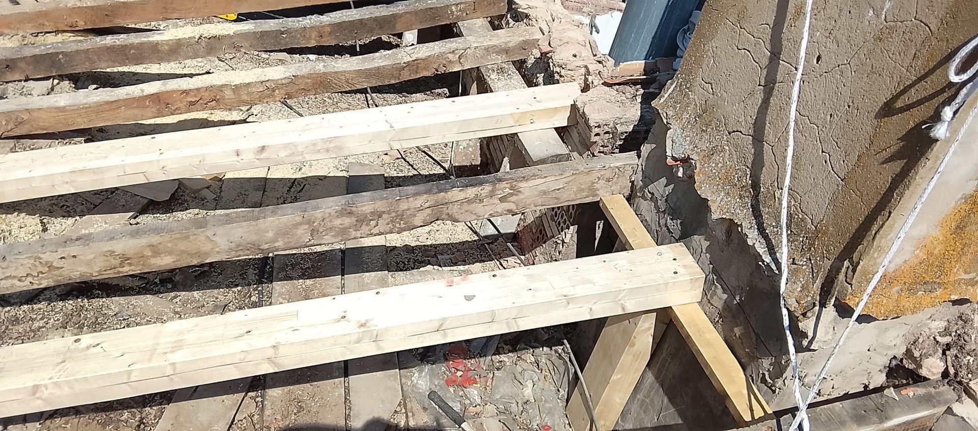 Vigas de madera colocadas sobre unos cimientos de tierra, junto a un muro de hormigón y tuberías de servicios públicos