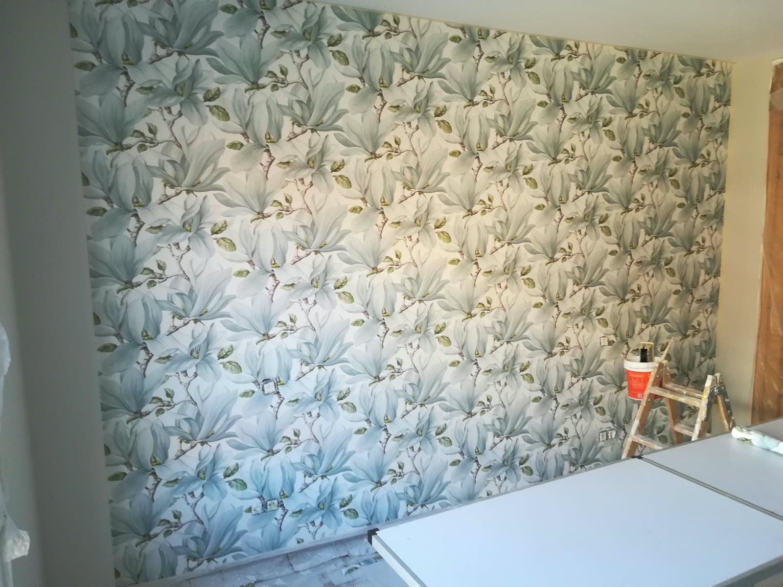 Se está colocando papel pintado con motivos florales en una habitación, con una escalera y materiales de pintura cerca