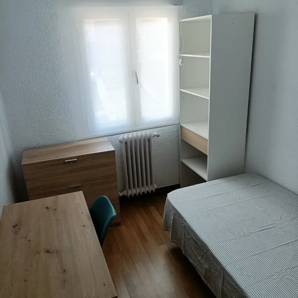 Habitación pequeña con cama, escritorio, silla, estantería, cómoda y ventana con radiador.