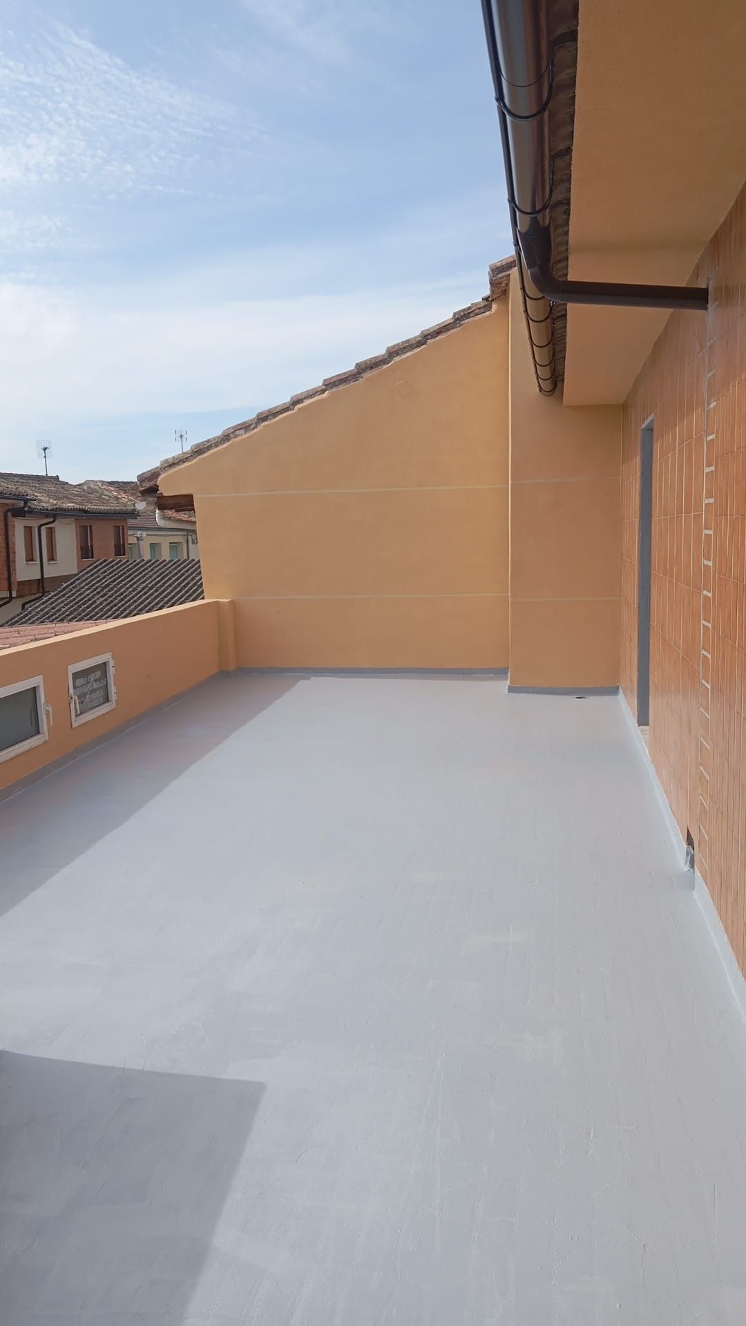 Terraza vacía en la azotea, con paredes beige, suelo blanco y edificios vecinos bajo un cielo azul