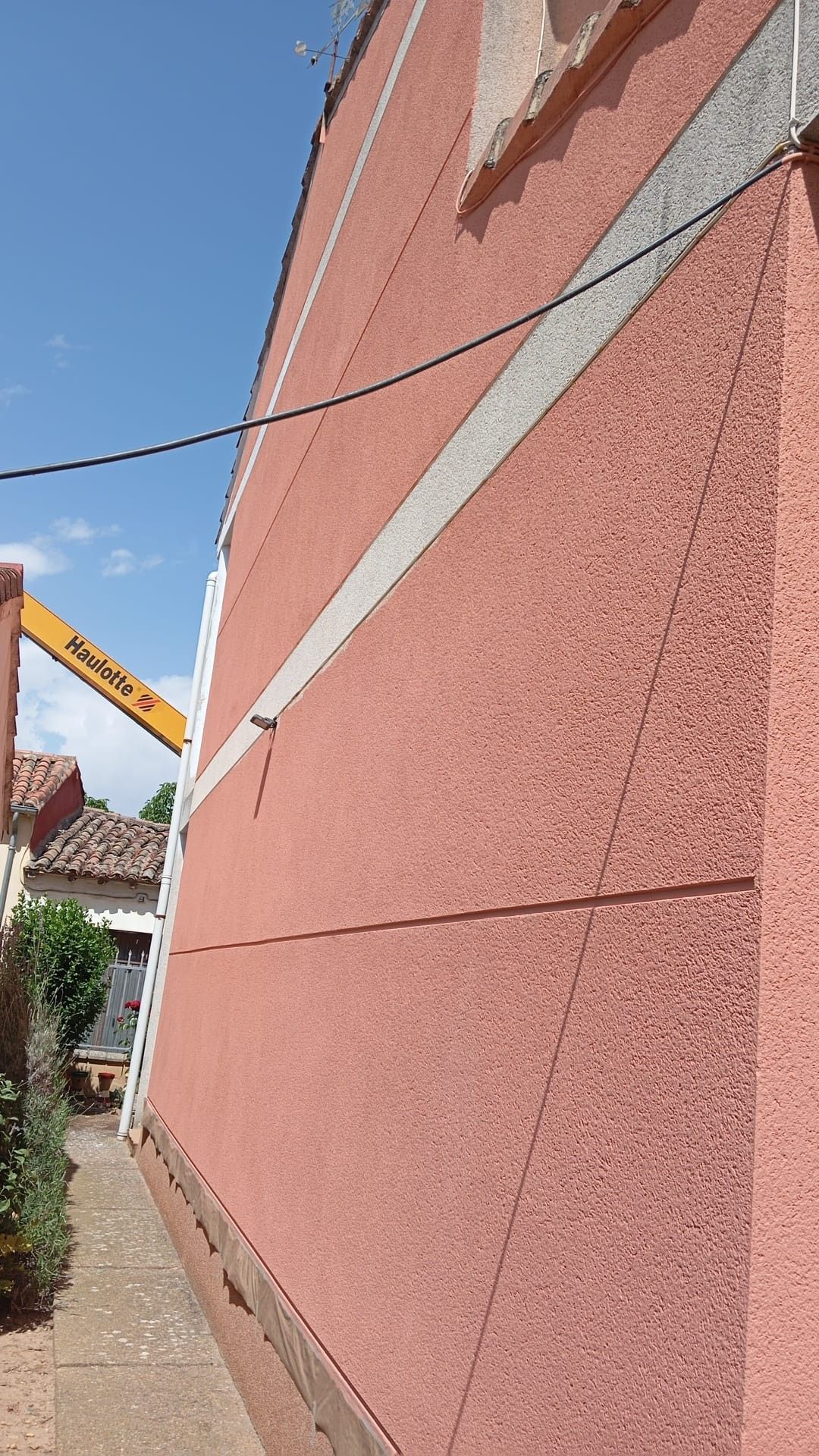 Pared de un edificio de estuco rosa con una franja gris diagonal junto a una acera estrecha bajo un cielo azul
