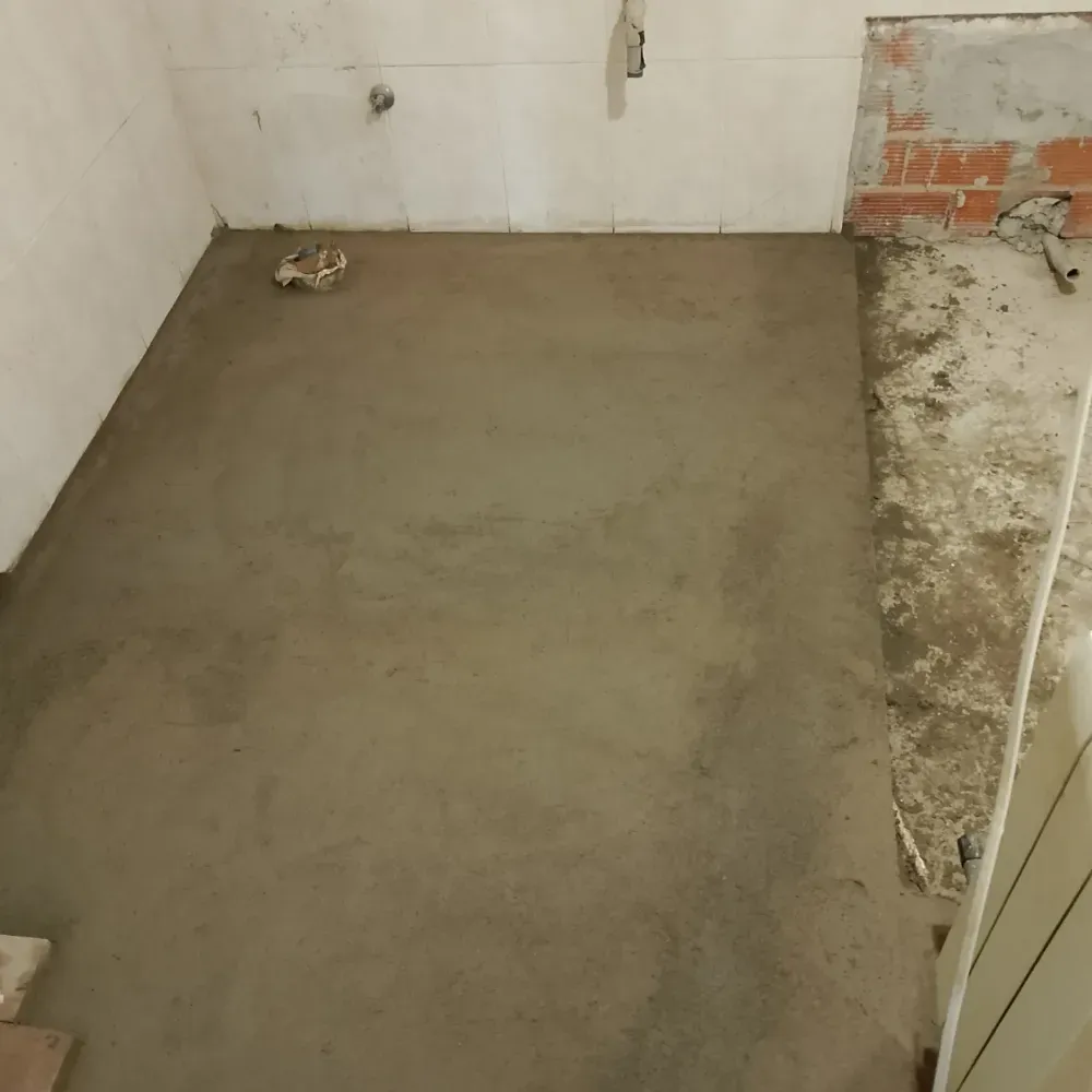 Habitación pequeña sin terminar, con suelo de hormigón húmedo y paredes de ladrillo visto y azulejos.