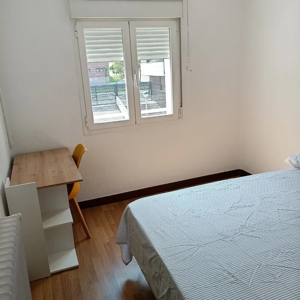 Habitación pequeña con cama, escritorio, silla y una ventana que deja entrar la luz natural.