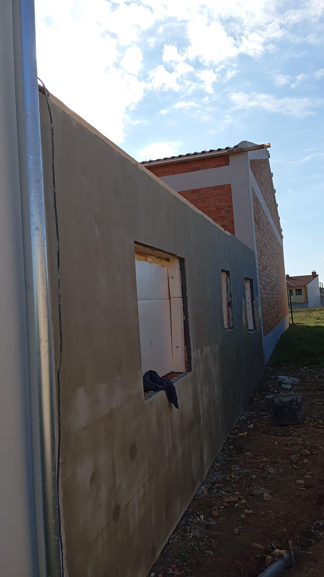 Callejón estrecho junto a una pared de estuco gris con una ventana pequeña, que conduce a una casa bajo un cielo azul.