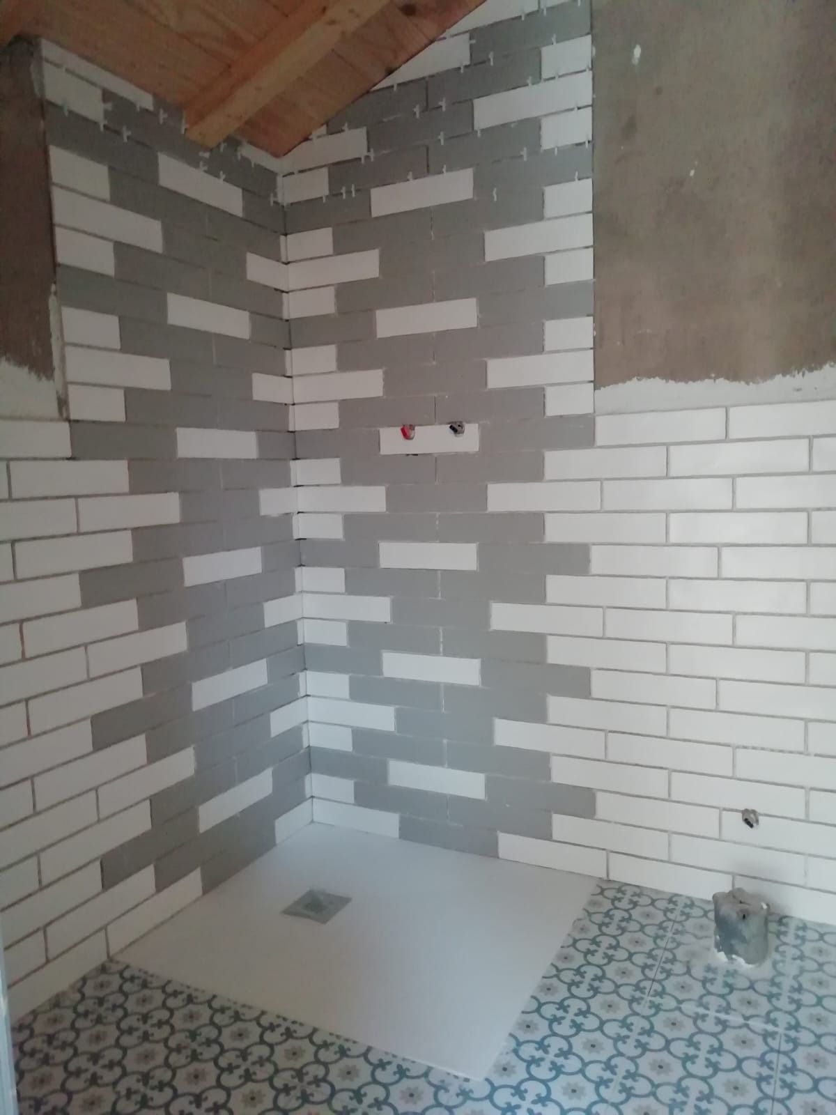 Esquina de ducha de baño parcialmente alicatada, con plato de ducha blanco y paredes con azulejos blancos y grises en patrón de ladrillo