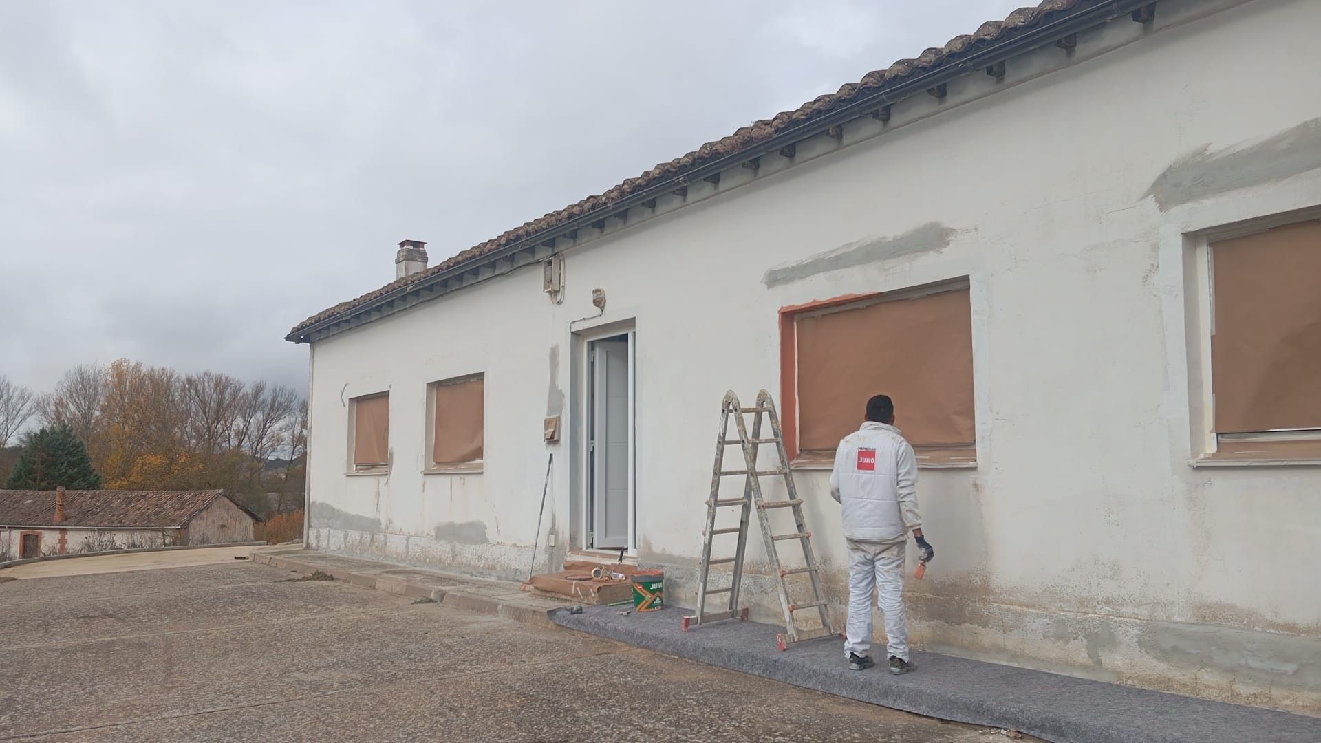 Un trabajador pintando la pared exterior de una casa blanca, con una escalera y herramientas en un camino de grava.